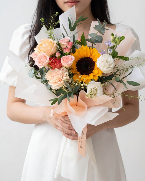 Bright Smile — a sunflower with peach and white roses, carnations and greenery, cream paper wrap, held by a model
