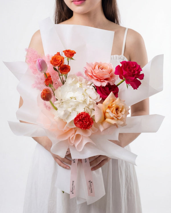 Palm Springs — white hydrangea, orange spray roses, a tulip, pink carnation and red rose in white wrap, held by a model.