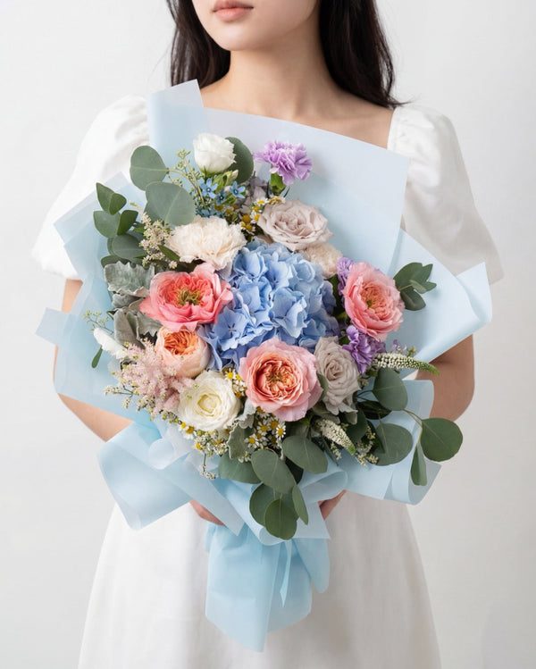 Sweetest Romance — blue hydrangea with coral garden roses and white ranunculus, blue paper wrap, held by a model