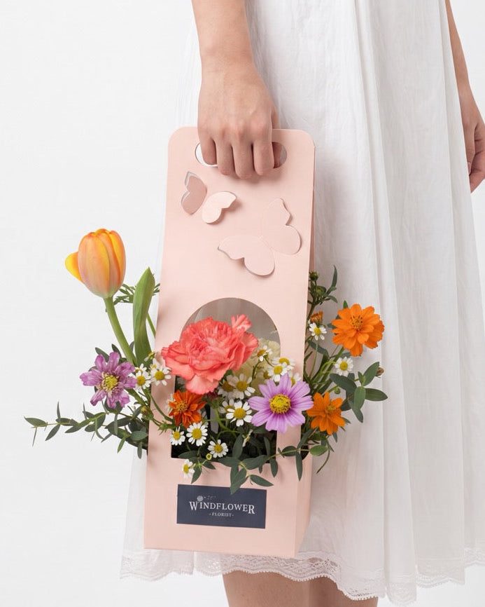 Blush Hour — orange tulip, coral carnation and purple flowers in a blush pink butterfly gift box, held by a model