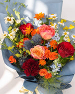 Cosmic Cheers — orange cosmos, red roses and blue thistle with white daisies wrapped in dusty blue paper, close-up