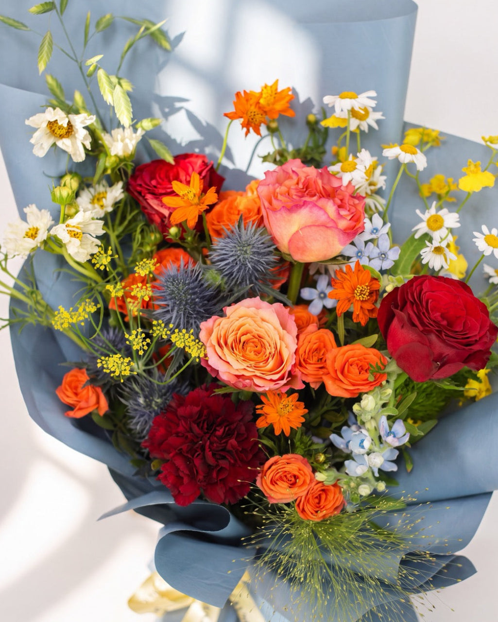 Cosmic Cheers — orange cosmos, red roses and blue thistle with white daisies wrapped in dusty blue paper, close-up