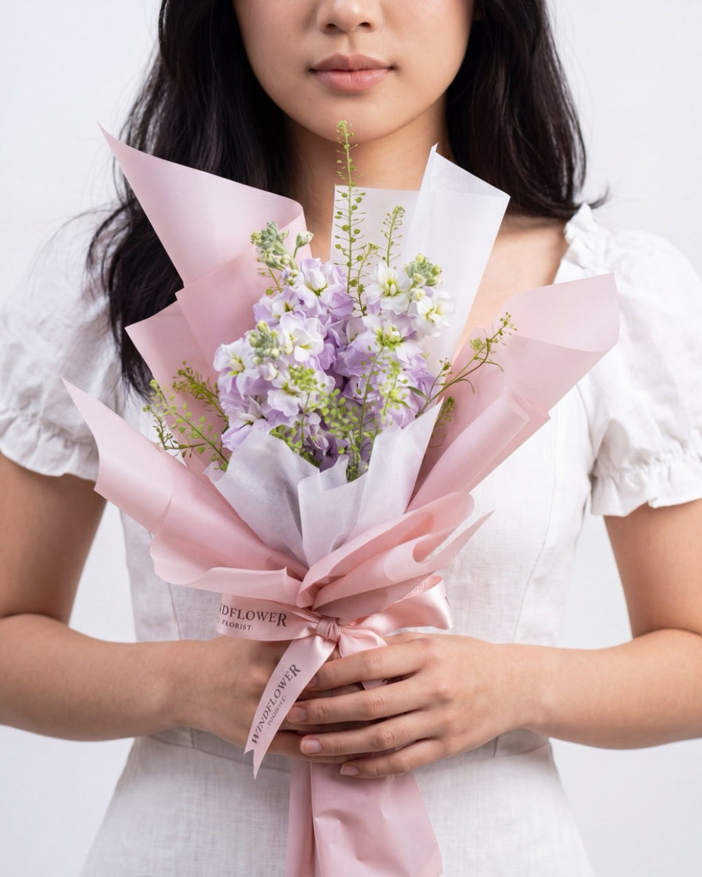 Trio Matthiola In Lilac — lilac stock flowers with greenery, wrapped in white and pink paper, held by a model
