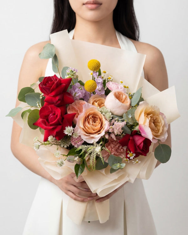 Koibito — red roses, peach garden roses, craspedia and lilac stock in cream wrap, held by a model.