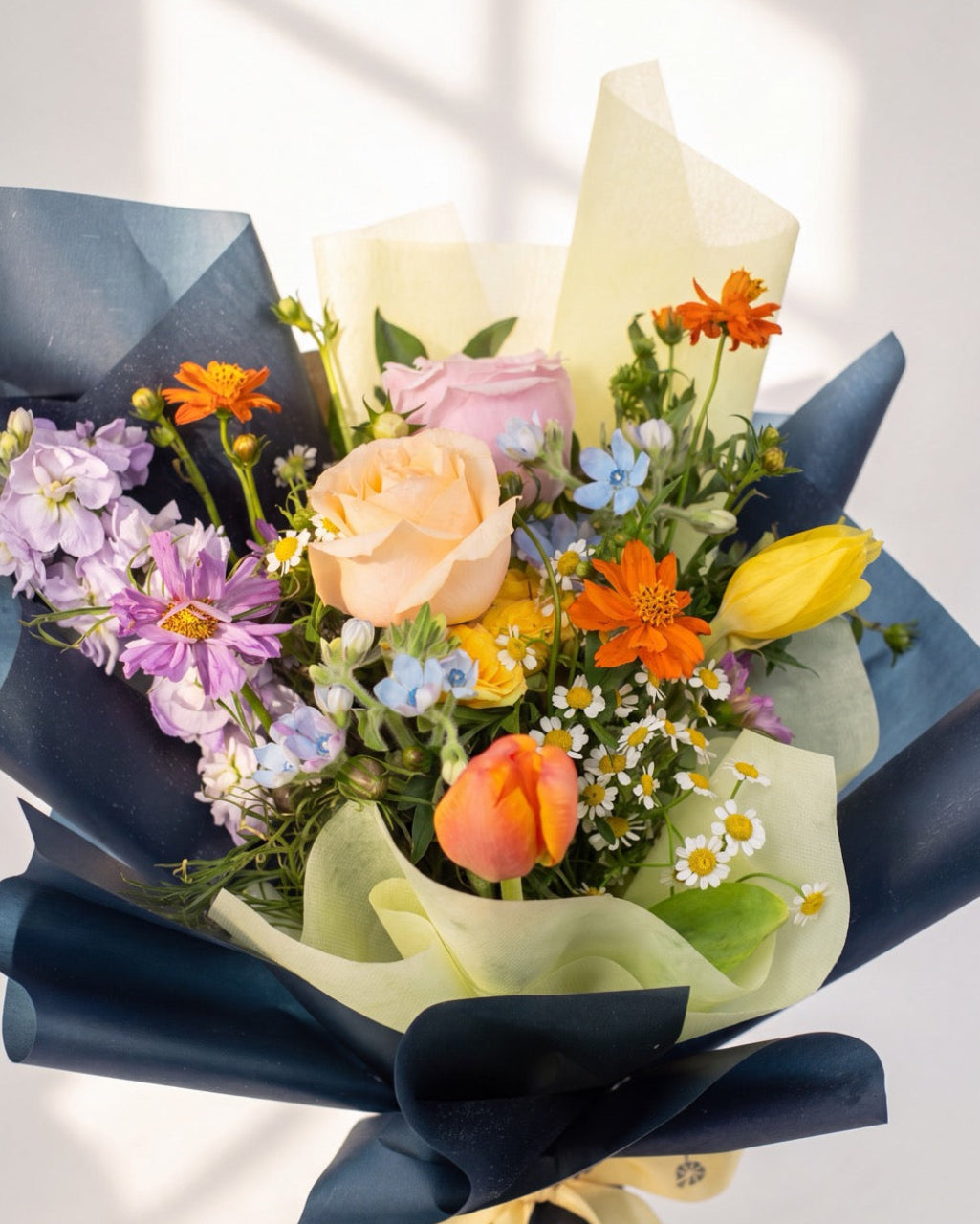 Soft Glow — peach and pink roses with purple stock and orange cosmos wrapped in navy and cream paper, close-up