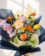 Soft Glow — peach and pink roses with purple stock and orange cosmos wrapped in navy and cream paper, close-up