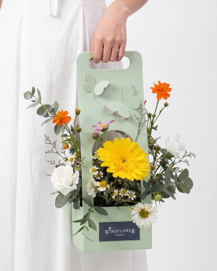Sunny Side Up — yellow gerbera daisy, orange cosmos and white roses in a sage green butterfly gift box, held by a model