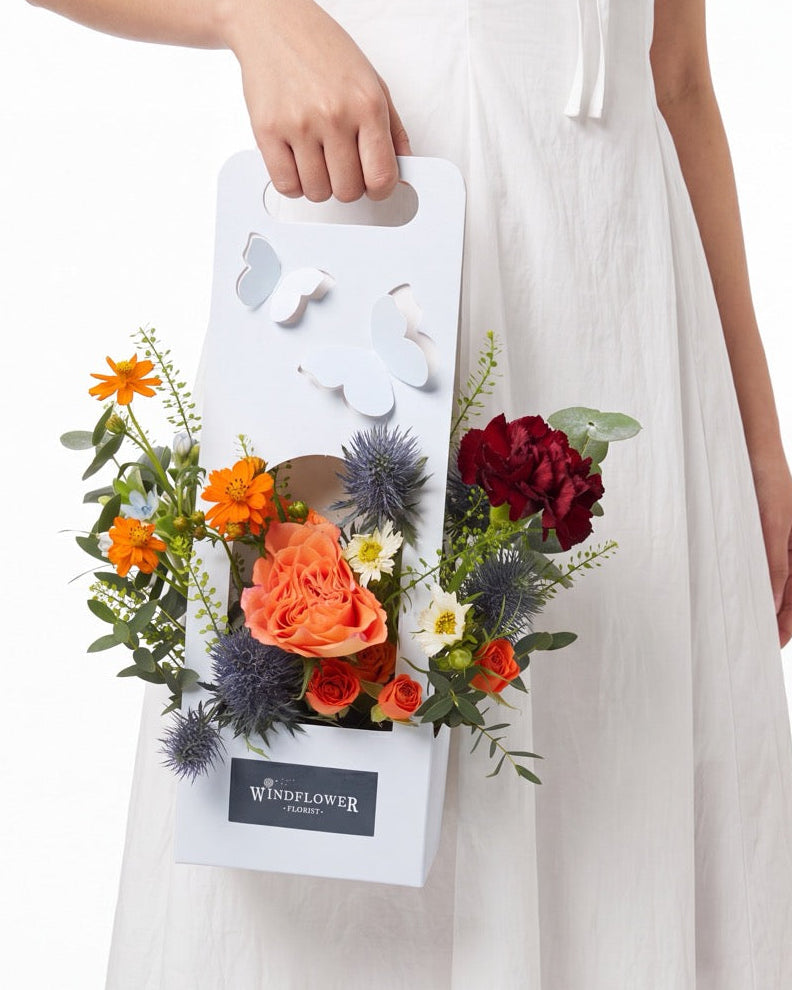 Tiny Garden Club — orange cosmos, coral rose and blue thistle with red carnation in a white butterfly gift box, held by a model