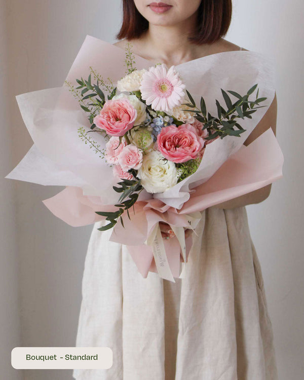 Mikoto — pink garden roses, a pink gerbera daisy and white ranunculus with greenery, pink paper wrap, held by a model